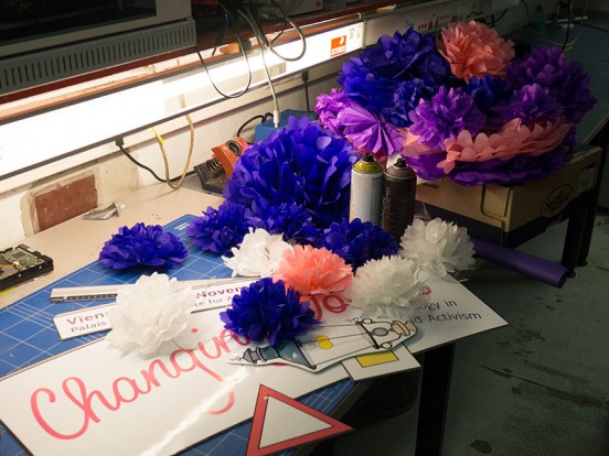 The picture shows a workbench with cutting mats, covered in colourful paper decorations. The decorations are big pink, purple and white tissue paper puffs in different sizes as well as big colourful printouts of the conference logo and poster artwork.
