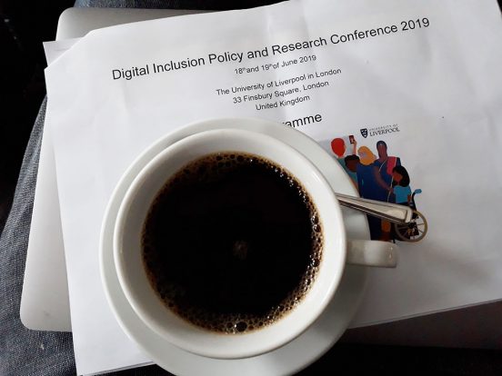A cup of coffee sits on top of a stack of papers, a laptop and the programme for the DIPRC conference on Angela's lap.