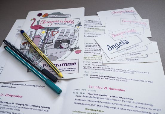 A few programmes and nametags (with pre-printed pronoun options) arranged on a desk with some pencils